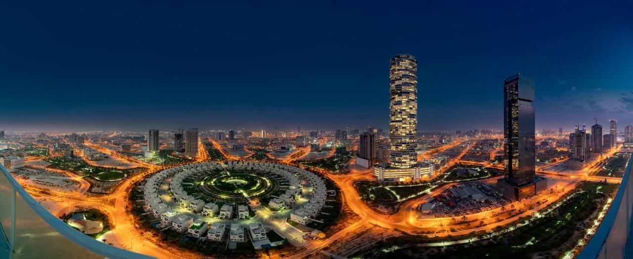 Five Jumeirah Village Dubai Buitenkant foto The photo showcases a cityscape illuminated at night. In the foreground, there is a circular arrangement of buildings or houses, creating a visually striking pattern. To the right, a tall, modern skyscraper stands out, featuring a sleek design with n