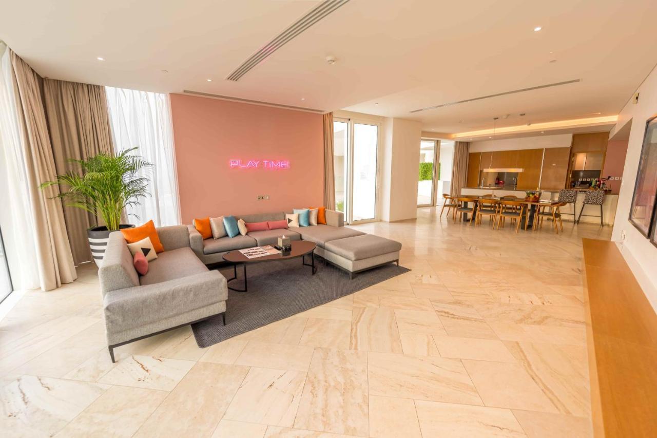 Five Jumeirah Village Dubai Buitenkant foto The photo shows a spacious and modern living area. There is a large, light-colored sectional sofa with colorful cushions, positioned on a dark rug. The wall behind the sofa is painted in a soft pink shade, and there is a subtle decorative element or 