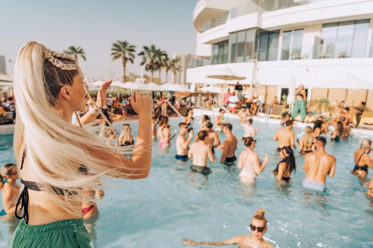 Five Jumeirah Village Dubai Buitenkant foto The photo features a lively pool party scene. In the foreground, a woman with long, blonde hair is seen raising her arm as if enjoying the moment. The pool is filled with many people, some swimming and others socializing. The atmosphere appears festi