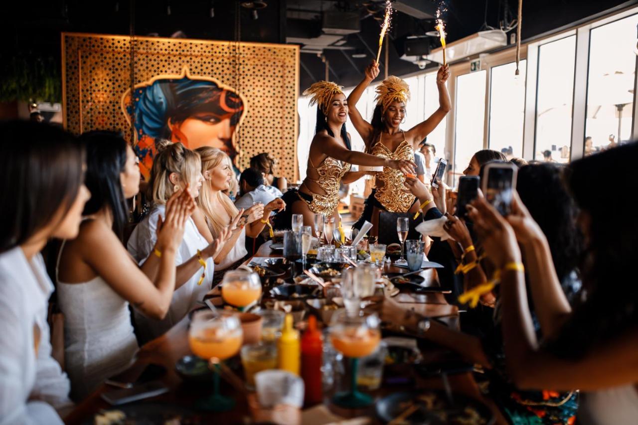 Five Jumeirah Village Dubai Buitenkant foto The photo depicts a lively restaurant scene where two performers are entertaining a group of seated diners. The performers, dressed in colorful and elaborate outfits, are holding sparklers, adding a festive atmosphere to the occasion. The diners, mos