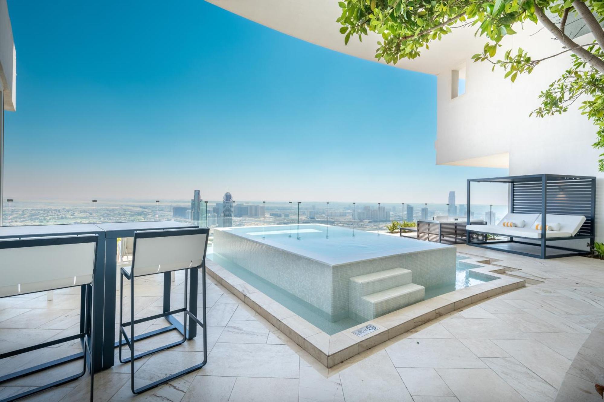 Five Jumeirah Village Dubai Buitenkant foto The photo shows a modern outdoor space featuring a sleek infinity pool. The pool appears to be situated on a terrace with a stunning view of a city skyline or landscape in the background. There are elegant lounge chairs and a shaded cabana area, crea