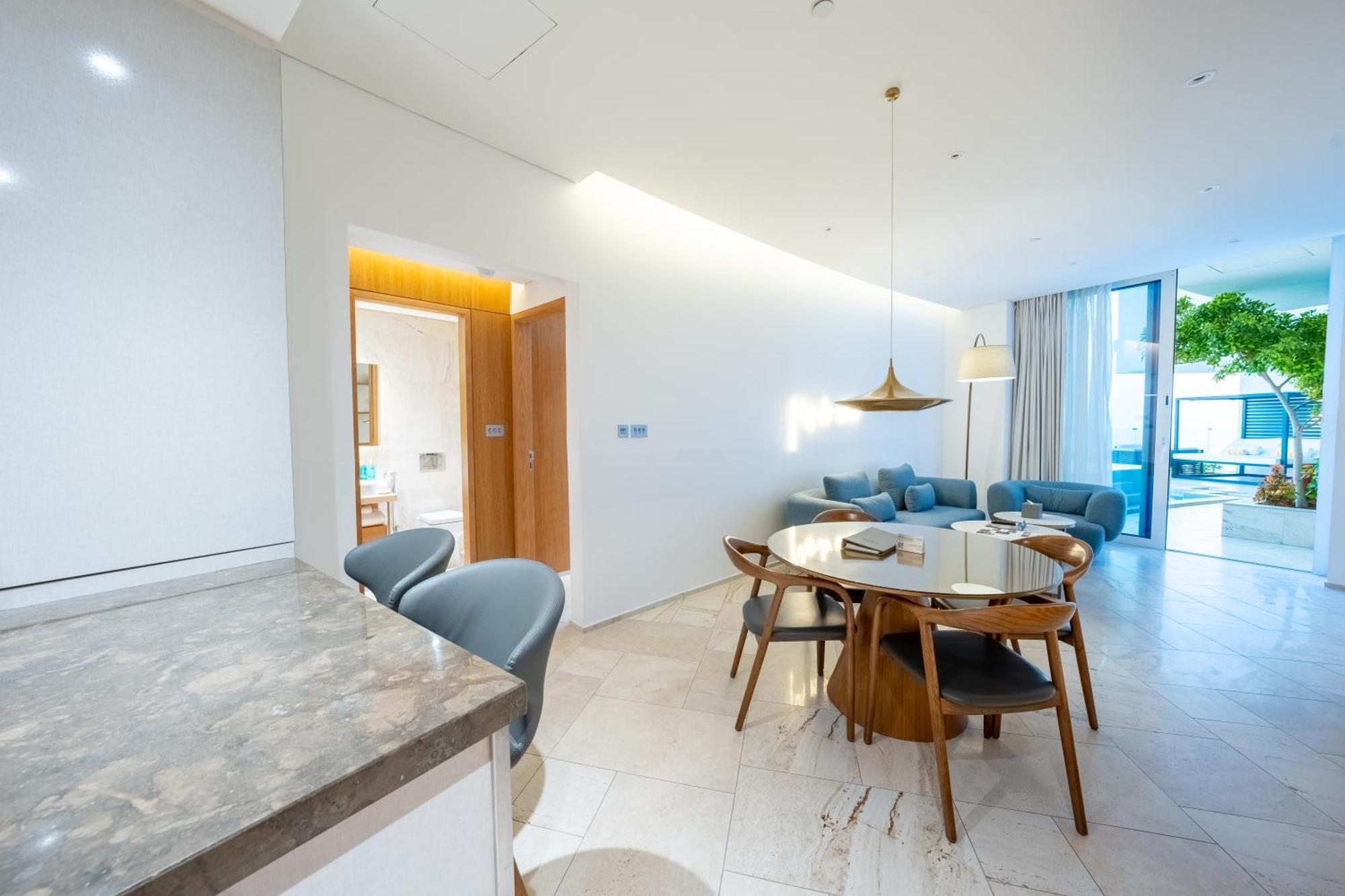 Five Jumeirah Village Dubai Buitenkant foto The photo shows a modern living space that combines a dining area and a lounging area. On the left, there is a kitchen counter with a marble surface and several gray chairs around it. In the center, there is a round dining table made of wood with sev