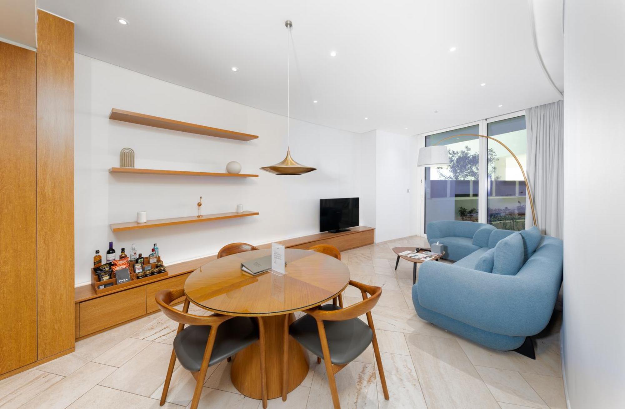 Five Jumeirah Village Dubai Buitenkant foto The image shows a modern living space that features a cozy and stylish design. On one side, there is a round wooden dining table surrounded by four chairs. The table is set with a small item, possibly a decorative piece or a book. 

A pendant light h