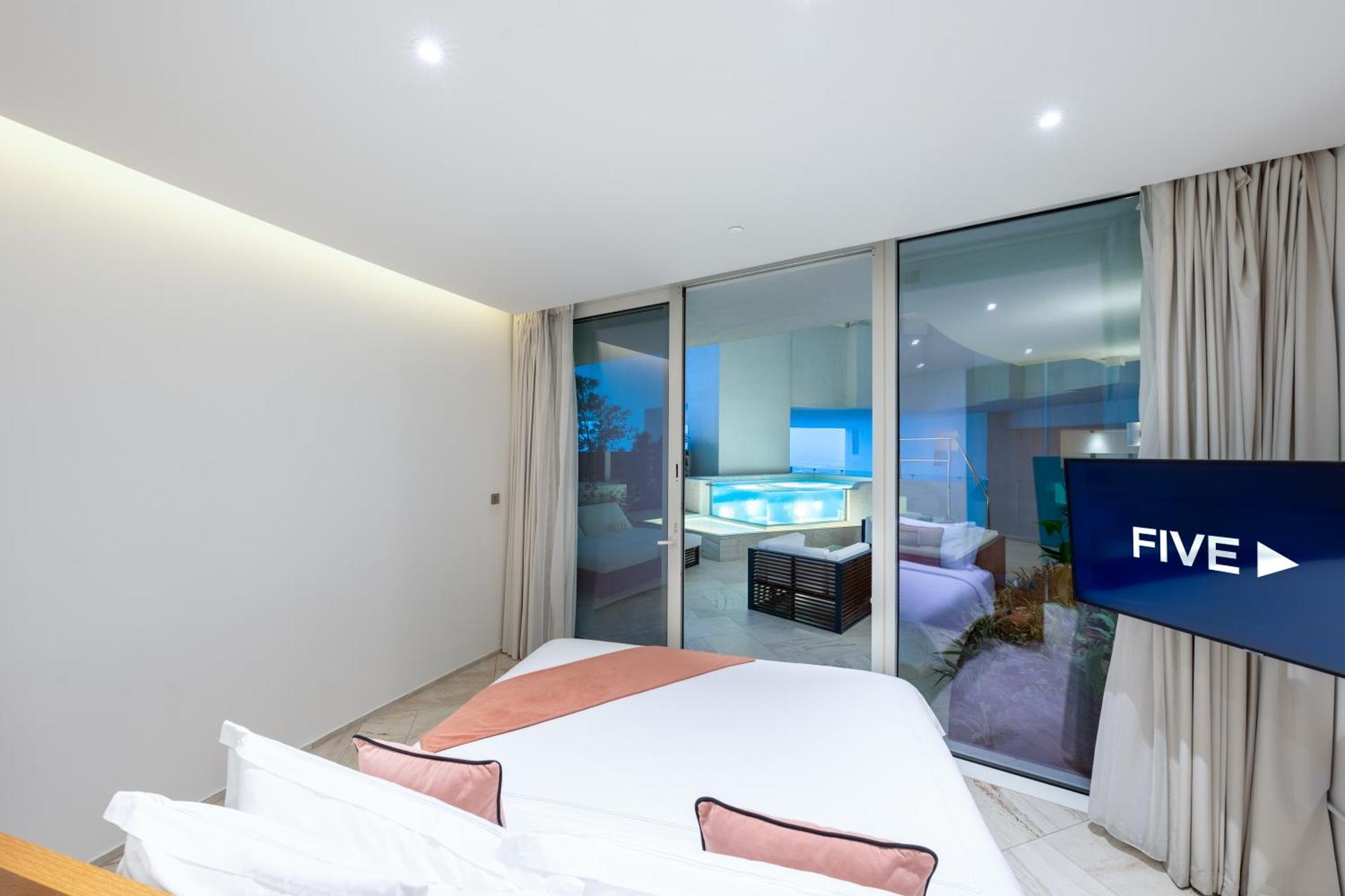 Five Jumeirah Village Dubai Buitenkant foto The photo shows a modern, stylish bedroom featuring a large bed with light-colored bedding and decorative pillows. There are large glass windows that open up to a view of an outdoor area, likely overlooking a pool or patio. The room has neutral-color