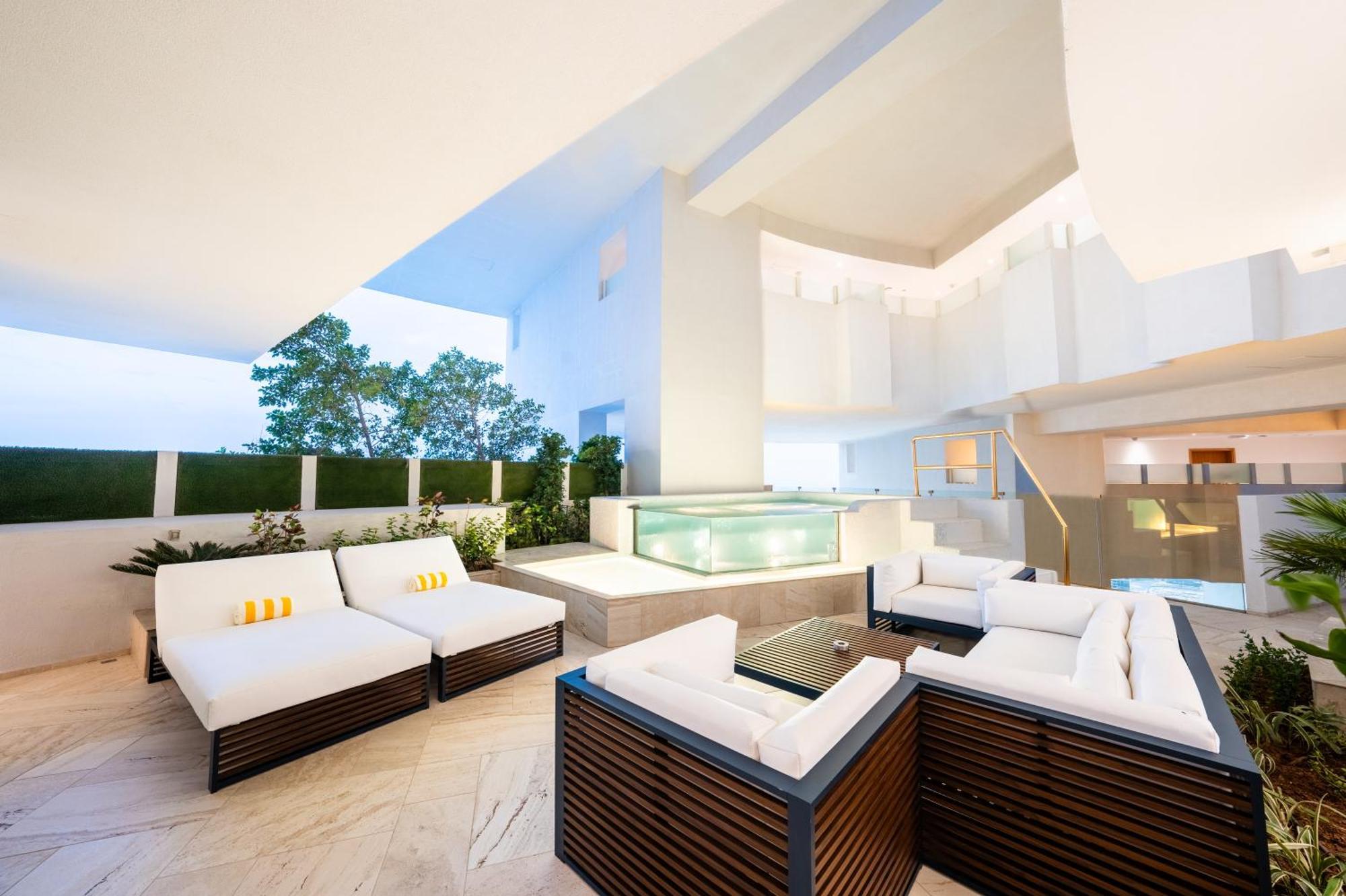 Five Jumeirah Village Dubai Buitenkant foto The photo depicts a modern outdoor lounge area featuring comfortable seating arrangements. There are several white sofas with dark wooden frames, arranged around a glass coffee table. In the background, there is a sleek water feature or pool, surroun