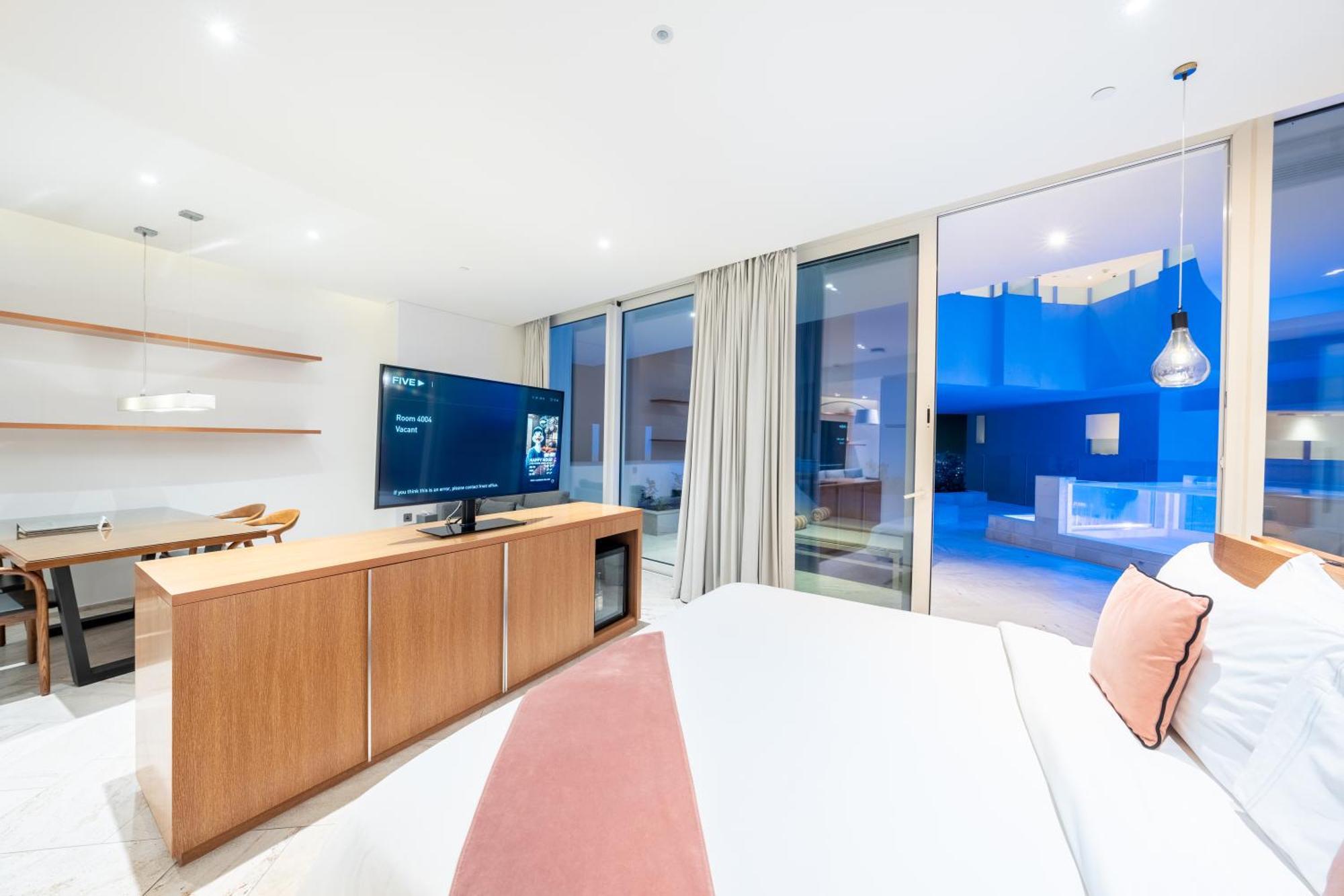 Five Jumeirah Village Dubai Buitenkant foto The photo depicts a modern hotel room with a clean and minimalist design. A large bed is shown, topped with white linens and decorative pillows. Opposite the bed, there is a wooden TV stand with a flat-screen television. The room features large windo
