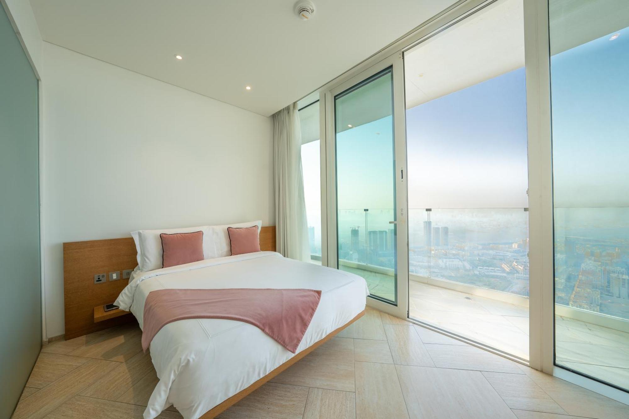 Five Jumeirah Village Dubai Buitenkant foto The photo shows a modern bedroom with a large bed featuring white bedding and accent pillows in a soft pink color. The room has floor-to-ceiling glass doors that lead out to a balcony, providing a panoramic view of the outside. The walls are painted 
