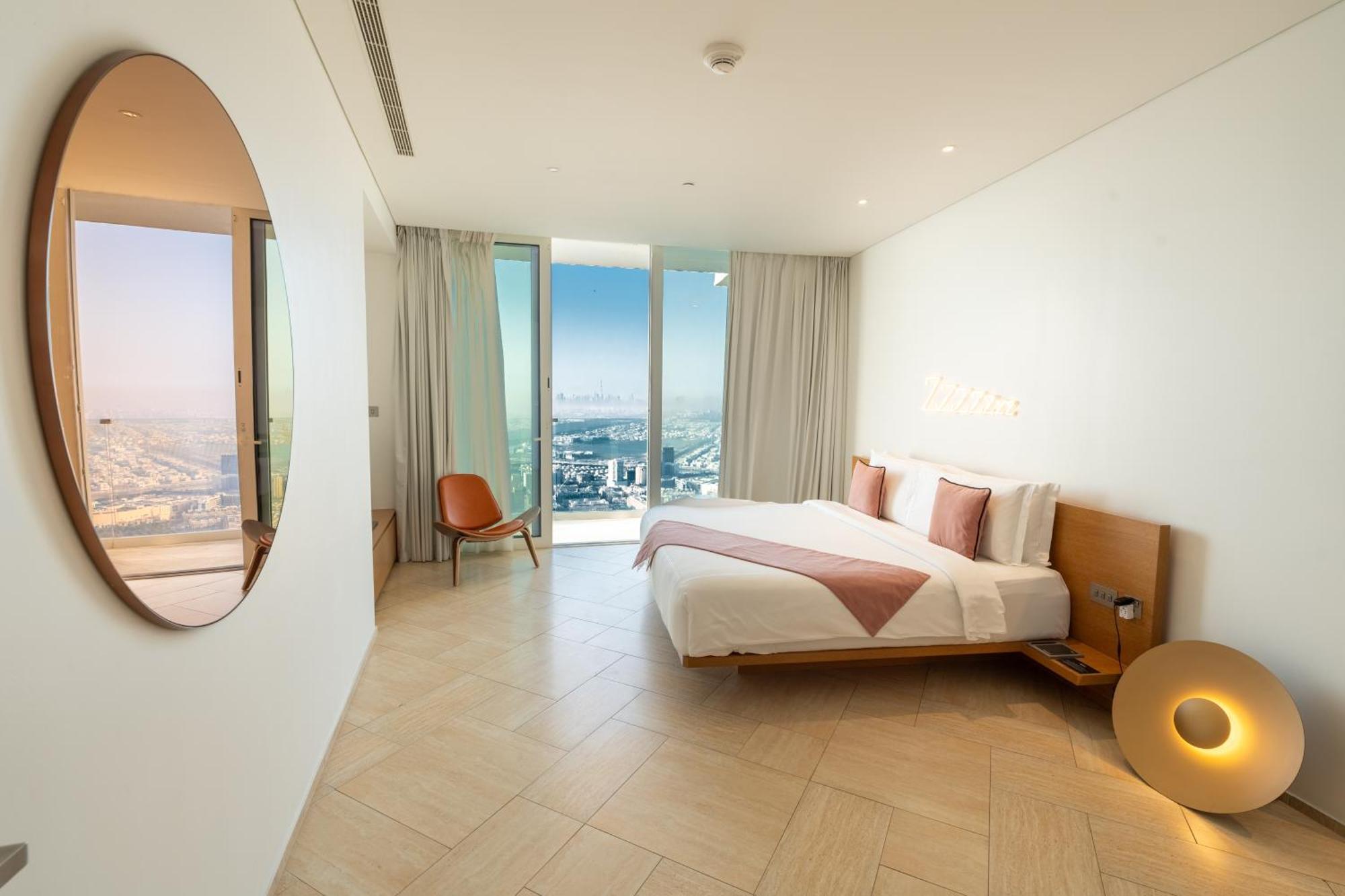 Five Jumeirah Village Dubai Buitenkant foto The photo shows a modern bedroom with a minimalist design. There is a large bed with white bedding and pink accents, positioned against a wall. To the side, there is a round mirror and a stylish chair. The room features large windows dressed with lig