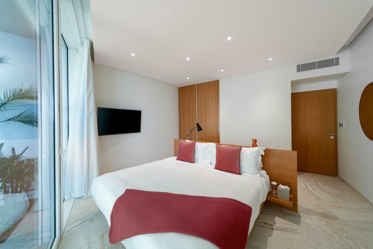 Five Jumeirah Village Dubai Buitenkant foto The photo shows a modern and stylish bedroom. It features a bed with white bedding and two red decorative pillows. There is a mounted flat-screen TV on the wall opposite the bed. The room has large glass windows that allow natural light to enter, and