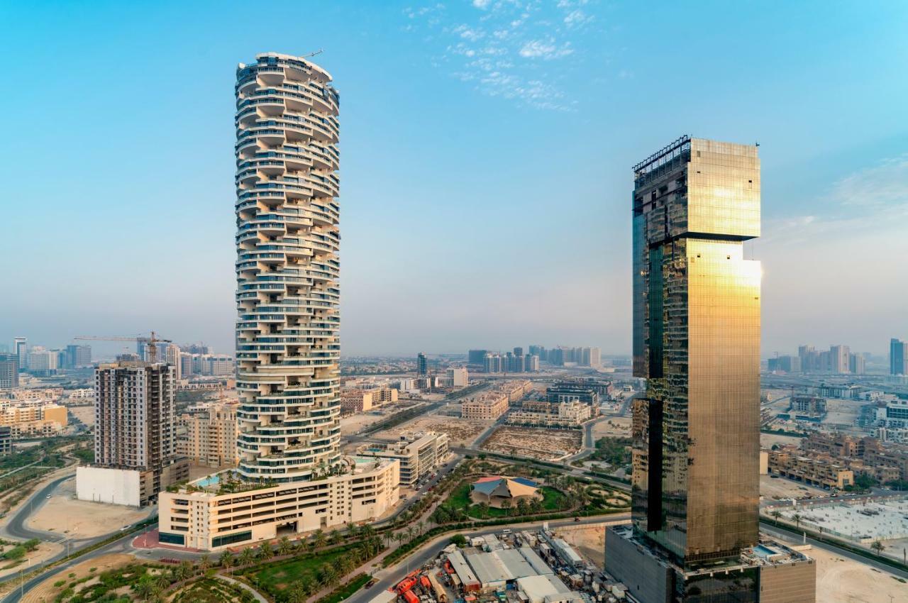 Five Jumeirah Village Dubai Buitenkant foto The photo shows two distinctive skyscrapers located in an urban area. On the left, there is a tall, cylindrical building with a unique, modern design characterized by its twisting, spiral shape and balconies at various levels. To the right, there is 