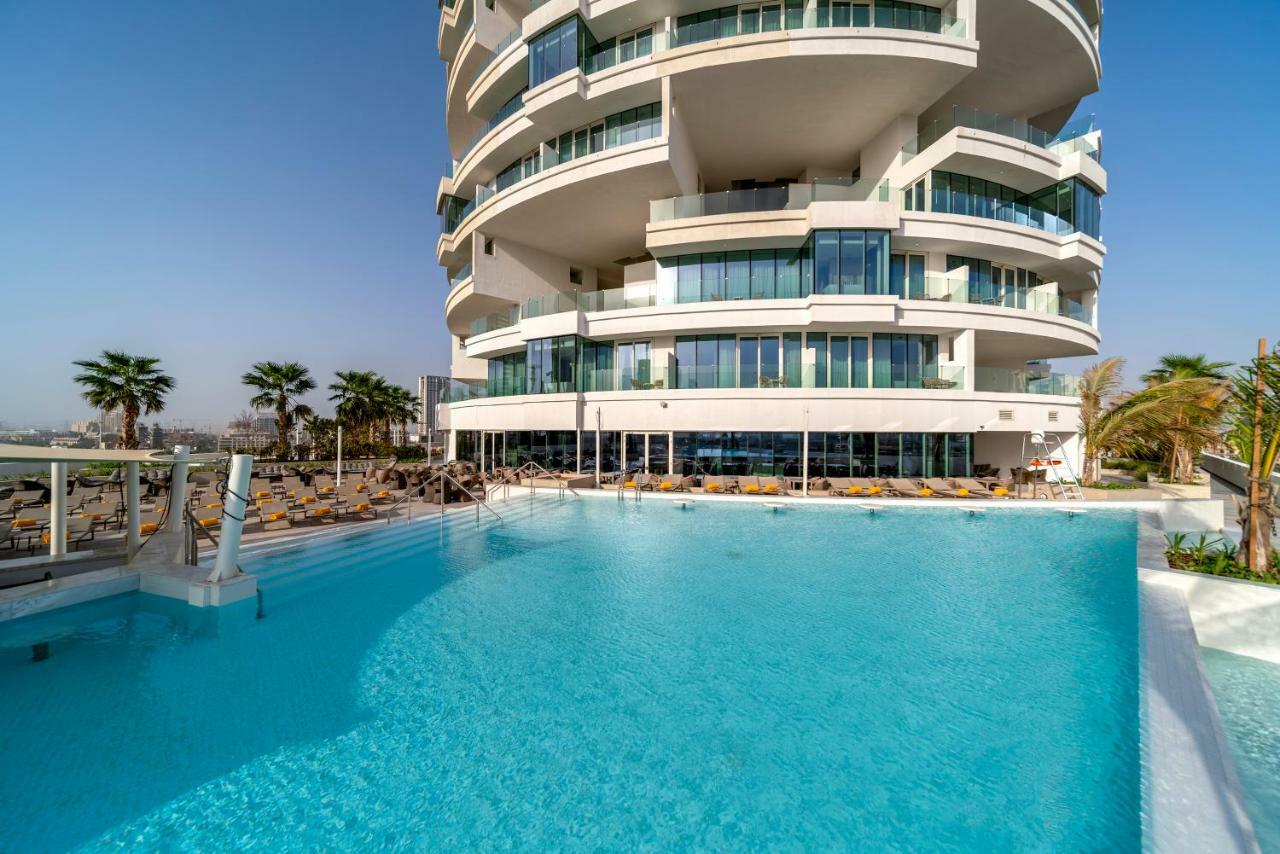 Five Jumeirah Village Dubai Buitenkant foto The photo depicts a modern building with a distinctive architectural design, featuring multiple curved balconies. In front of the building, there is a large, inviting swimming pool with clear, turquoise water. Surrounding the pool are loungers for re