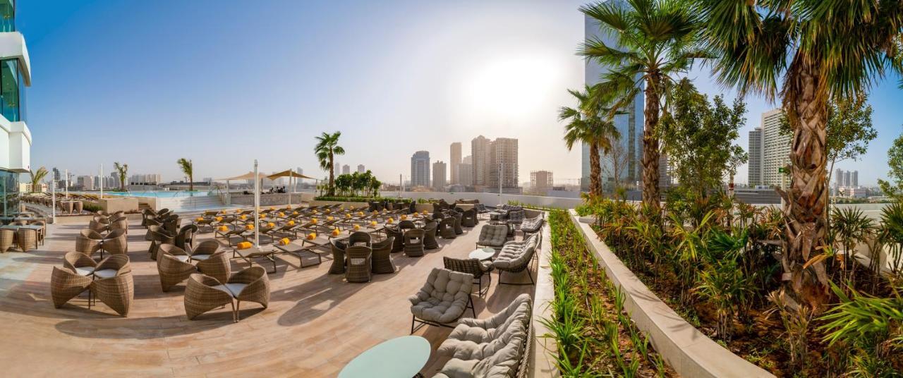 Five Jumeirah Village Dubai Buitenkant foto The photo showcases a luxurious outdoor seating area, likely part of a resort or hotel. In the foreground, there are well-arranged lounge chairs and seating options, some with cushions, placed on a tiled surface. The area is bordered by lush green pl